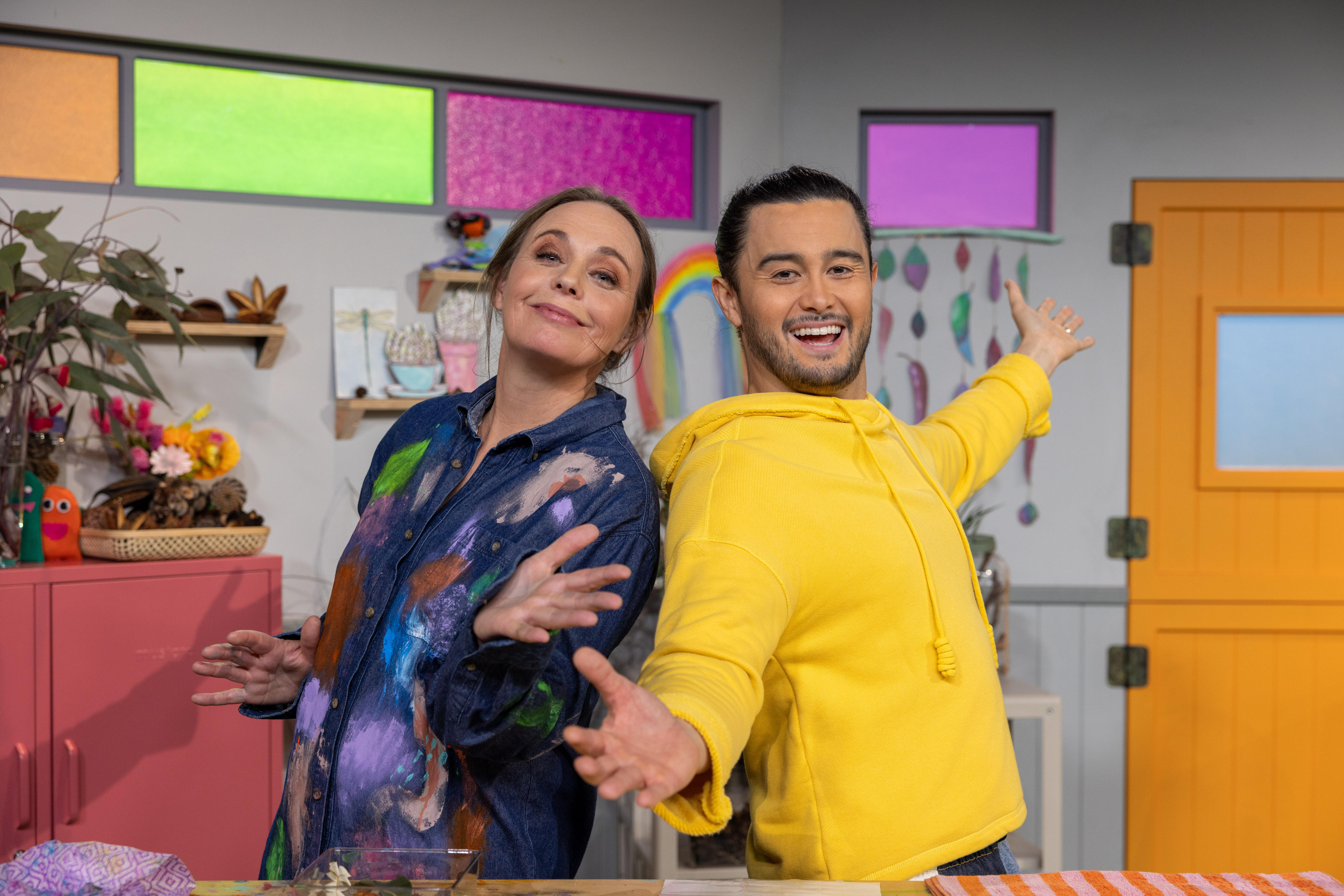 Two happy Play School presenters, one is dressed up in an art smocks in a playfully decorated art studio.