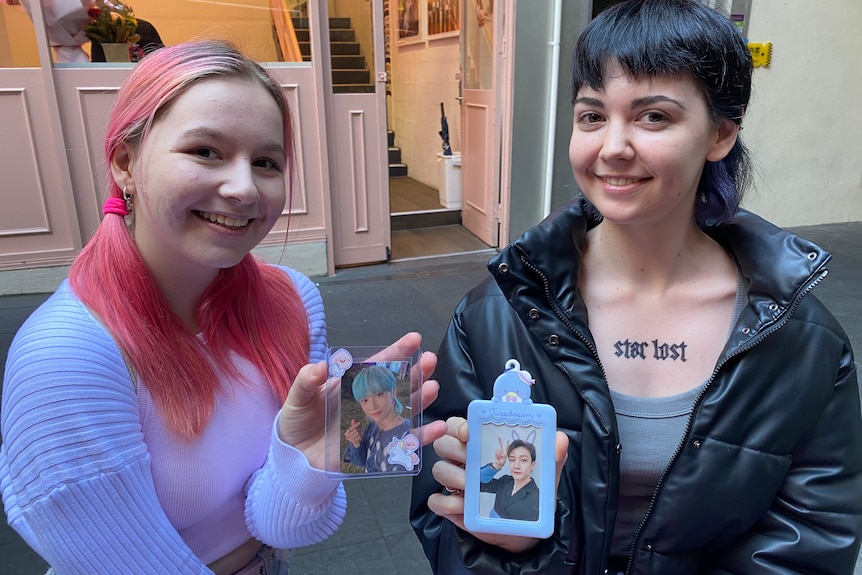 Two young women holding up photo cards of K-pop idols outside a store.