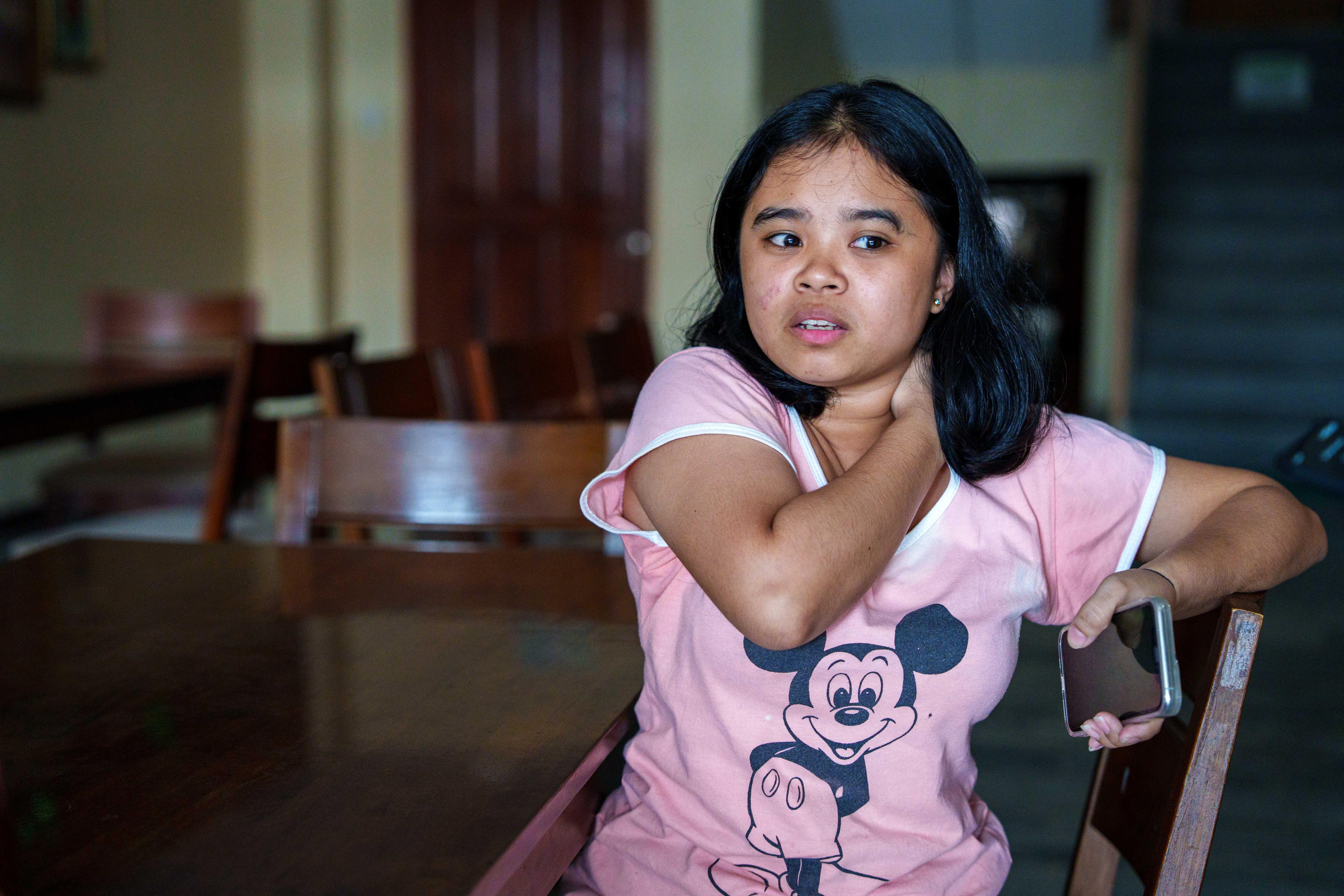 Uma jovem vestindo uma camisa rosa do Micky Mouse se apoia em uma cadeira enquanto segura um telefone.