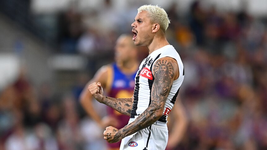 A Collingwood AFL player stands side-on to the camera, shouting in celebration as he pumps his fists.