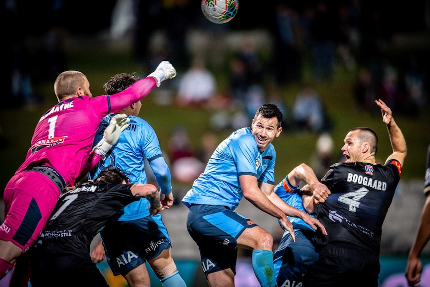 A goalkeeper flies over a pack to punch the ball as a number of players duck for cover or watch on