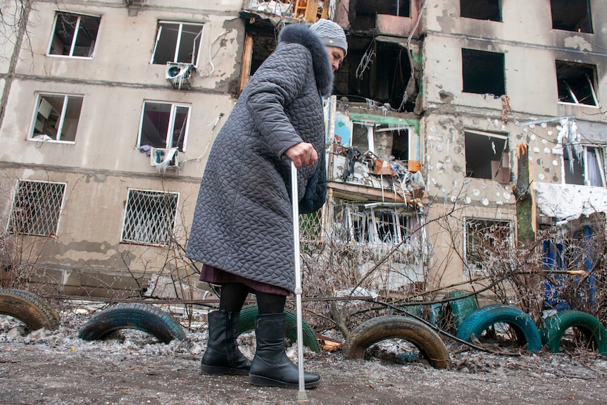 An elderly woman with a walking stick walks past a damaged apartment building, where rooms are exposed after being destroyed.