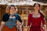 Two women in 1800s period-costume dresses walk past timber huts.