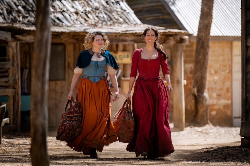 Two women in 1800s period-costume dresses walk past timber huts.