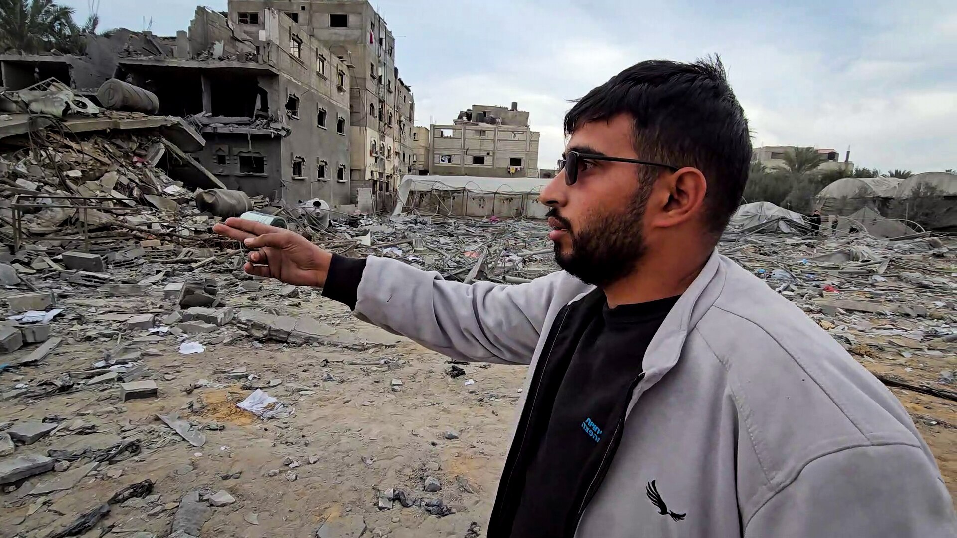 A man in grey zip-up hoodie points to piles of cement rubble