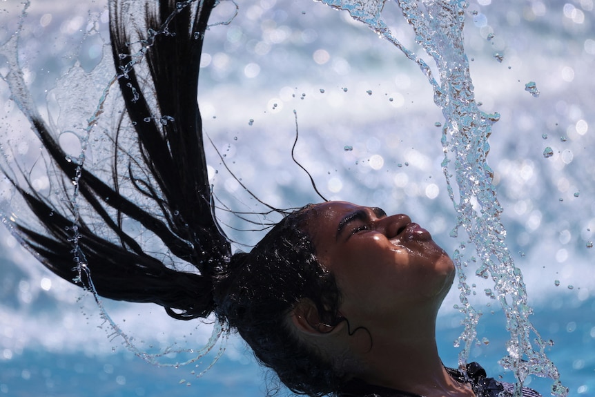 A girl sitting in water whips her long hair back causing a stream of water to drop from her wet hair