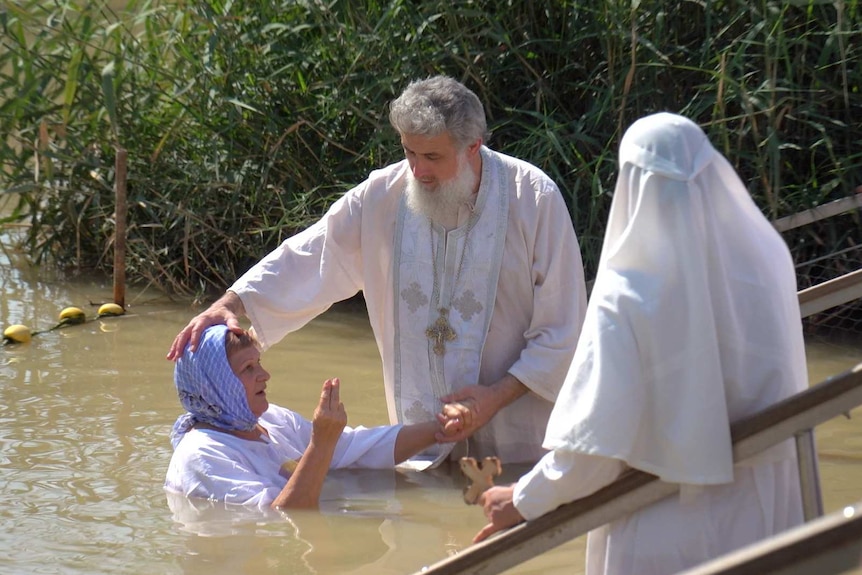 A man in a white robe holds a woman's head as she is submerged in a river as a woman in a white robe watches.
