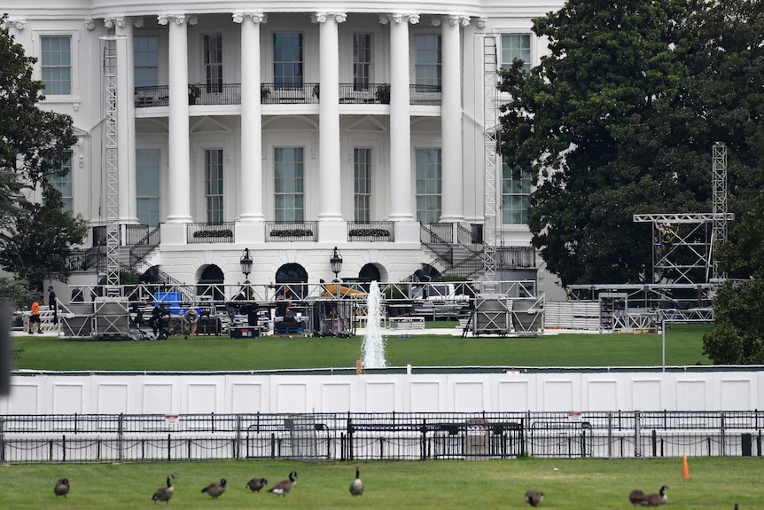 People set up poles and scaffolding on a green lawn in front of a white house.