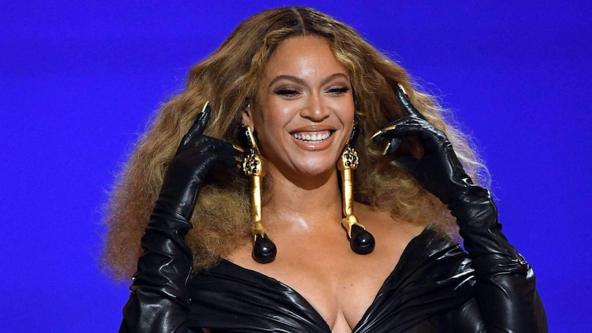 Beyoncé smiles against a blue backdrop wearing black gloves, a black dress and gold earrings
