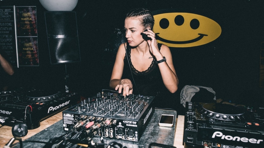 A woman holds headphones over her ears and stands at a DJ mixing desk