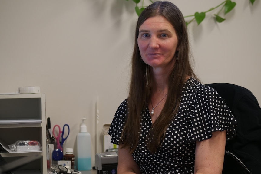 A woman with long brown hair and a black and white spotted top sits at a desk, looking at the camera.