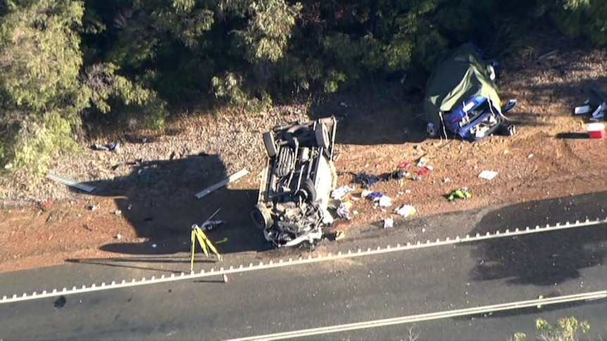 Two wrecked cars, one of them resting on its roof, and associated wreckage by the side of a road.