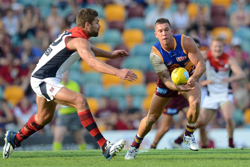 A Brisbane Lions AFL player looks to handball forward as a Melbourne defender closes in from the side.