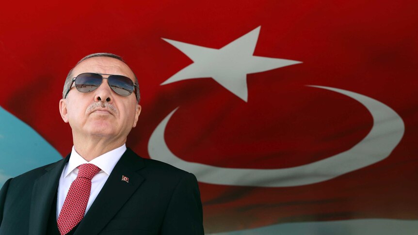 Turkish President Recep Tayyip Erdogan wears aviators and stands in front of the Turkish flag.