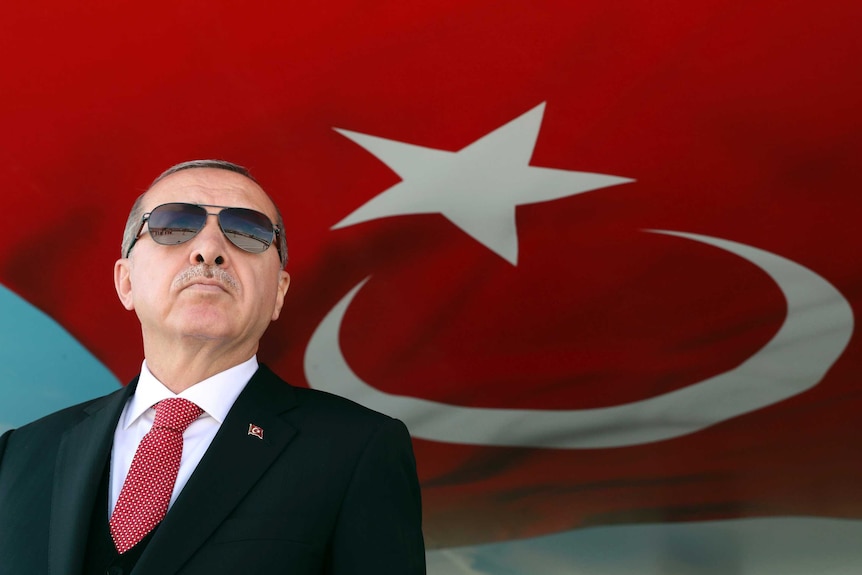 Turkish President Recep Tayyip Erdogan wears aviators and stands in front of the Turkish flag.