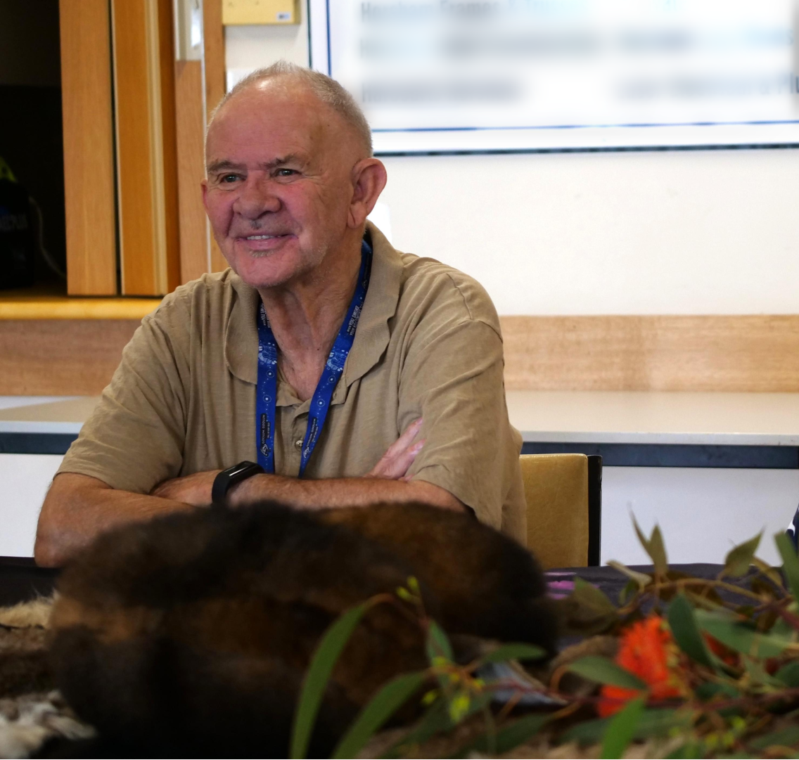 A man in a polo shirt and a blue lanyard sits smiling at a table with his arms crossed.