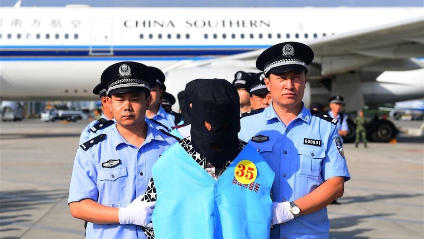 Chinese police stand with deported fraud suspects on the tarmac. The suspects are hooded and wearing numbered blue vests.