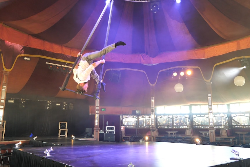 A man on a swing in a circus tent, balancing on his head.