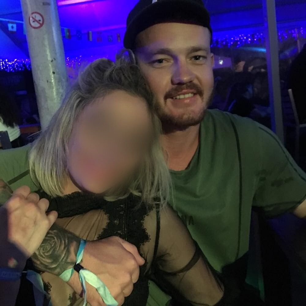 A man smiles next to a woman whose face has been blurred.