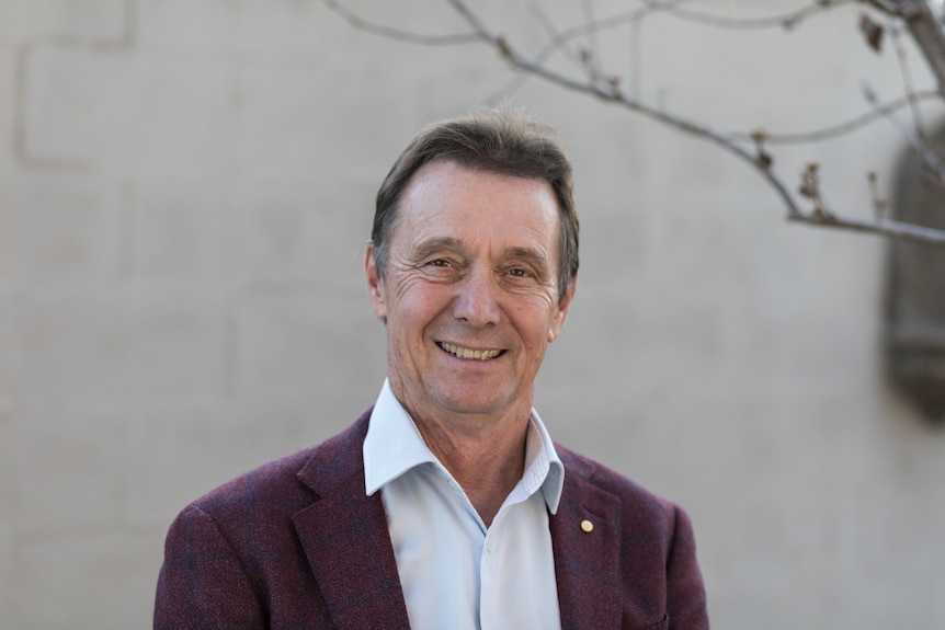 A smiling older Caucasian man in a purple blazer, light shirt, stands against a off-white wall, a branch visible in one corner.