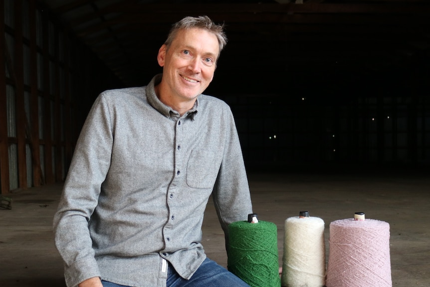 a man sits next to three large reels of spun wool