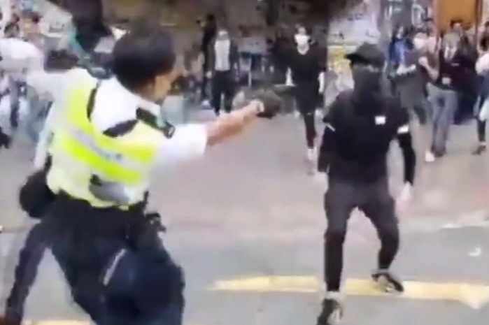 A grainy image from a social media video showing a police officer pointing a gun at a protester, who has their face covered.