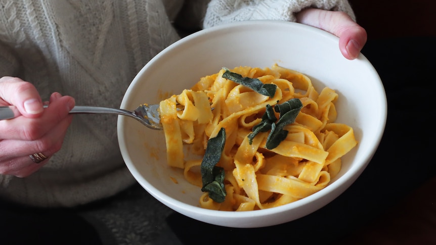 Close-up of person wearing knitted jumper and holding a bowl of creamy pumpkin pasta with buttered sage leaves.