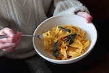 Close-up of person wearing knitted jumper and holding a bowl of creamy pumpkin pasta with buttered sage leaves.