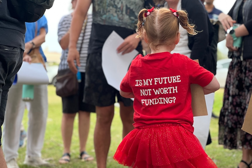 A close-up shot of a little girl from behind in a crowd of people, with her red shirt reading 'is my future not worth funding?'.