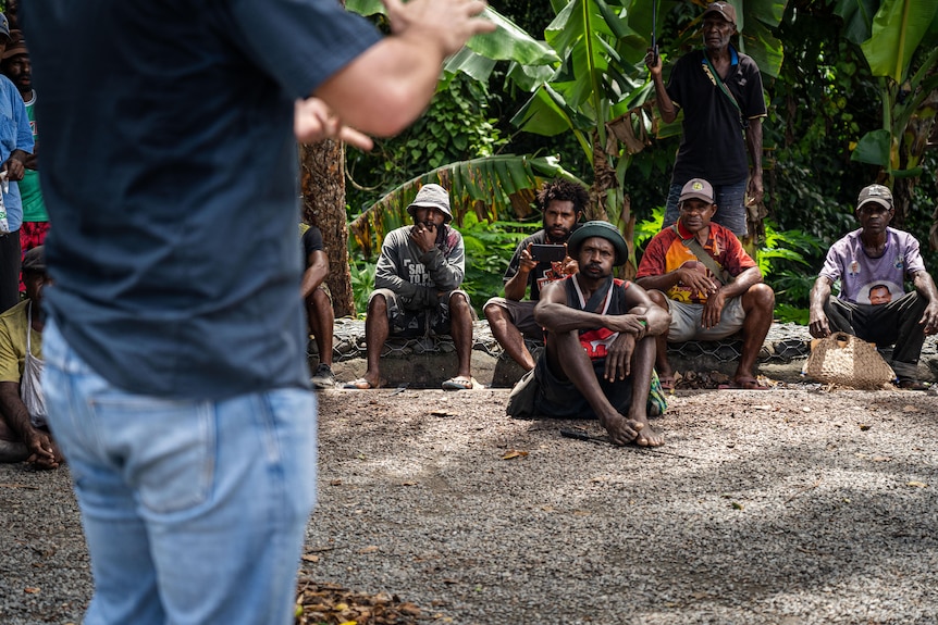 A group of peope sit on the ground surrounded by trees watching a man deliver a presentation.