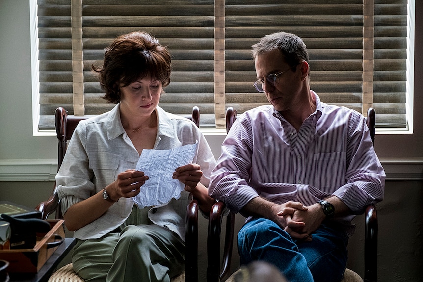 A woman with short brown hair, cream shirt sits and reads letter near window with closed shutters, a man sits next to her.