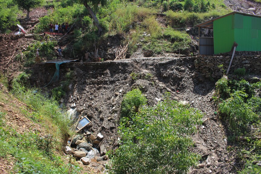 You view a wide photo of the aftermath of a landslide on a hill in daytime, with a house perched precariously over it.