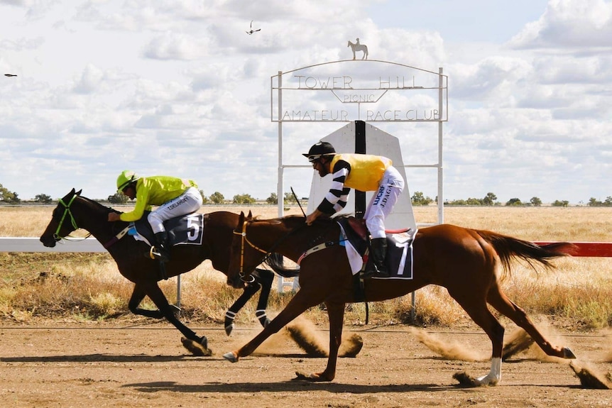 Two racehorse cross the finish line on red dirt race track, a large metal structure with a horse and rider on top in background.