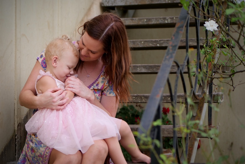 A small girl in a pink dress on a woman's lap sitting on stairs near a white rose bush.