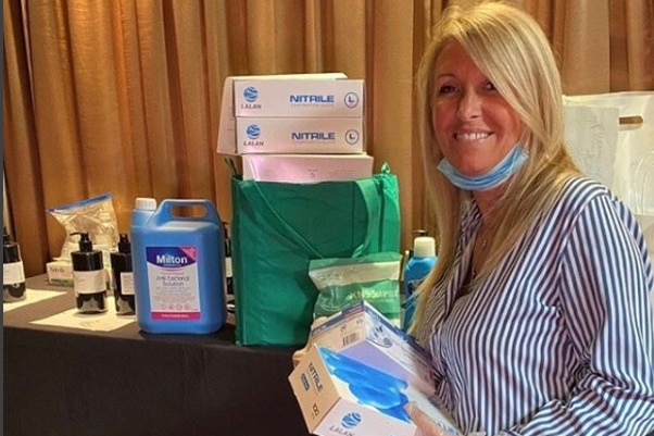 A woman with a surgical mask under her chin holds and stands in front of a variety of PPE and hand sanitiser.