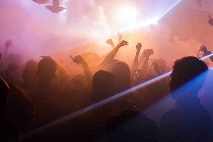 A group of dancers, dancing in a club with smoke machines and pink and yellow lights.