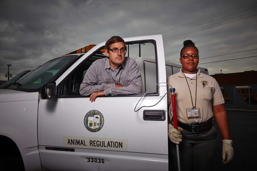 Louis Theroux stands leaning on the door of an animal regulation vehicle with an animal controller next to him