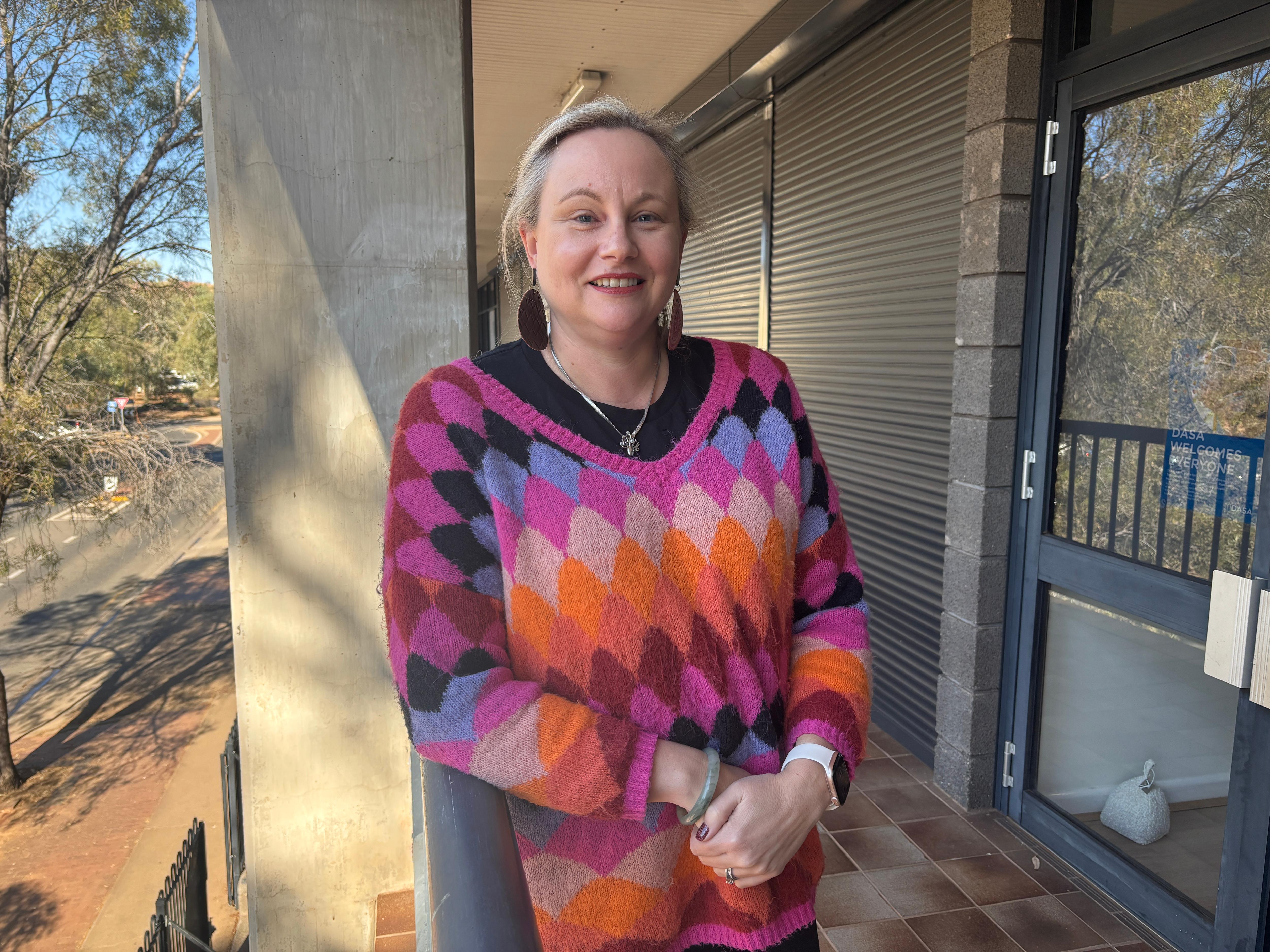 A woman wearing a bright pink and orange knit jumper leans against a railing on a balcony.