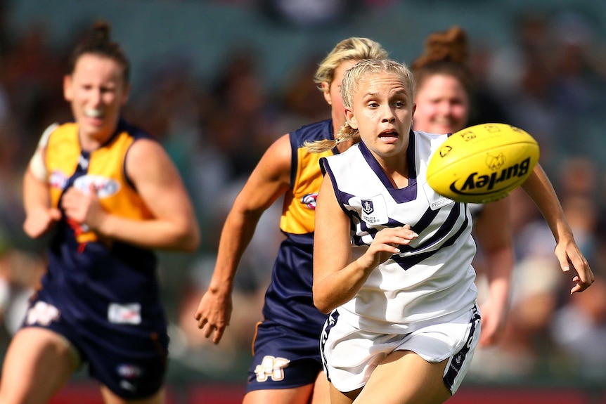 Fremantle Dockers player Courtney Ugle chases a loose yellow football as she is chased by two West Coast Eagles players.