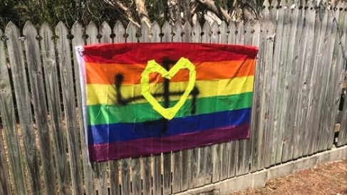 A rainbow flag on a fence is defaced with a spray-painted swastika.