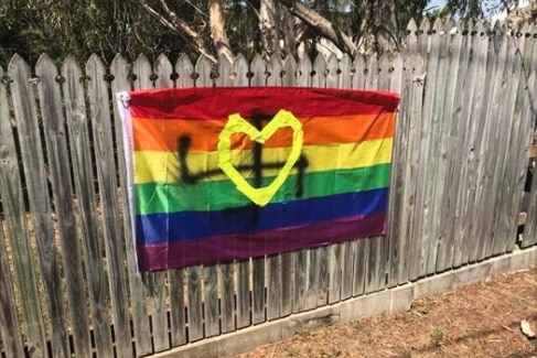 A rainbow flag on a fence is defaced with a spray-painted swastika.