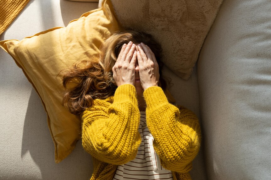 A woman laying down face up on a beige couch with yellow woolen cardigan, and hands covering her face.