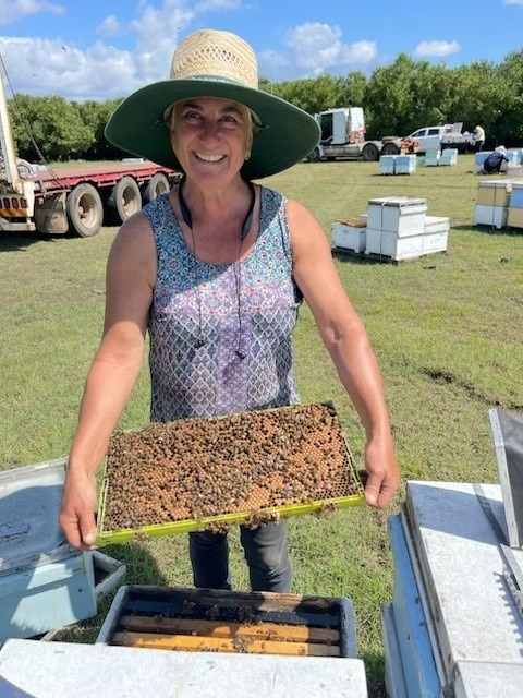 A woman in a broad-brimmed hat smiles with beehives in the background.