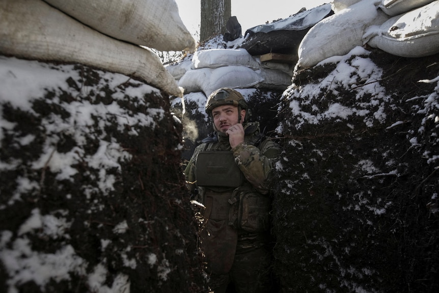 A man in military uniform holds a radio to his mouth. He stands surrounded by dirt walls dusted in snow