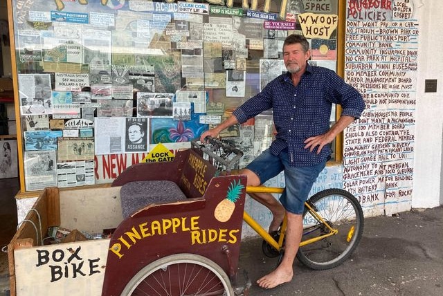 Bare-footed man on bike stands in front of shop that is covered in colourful text