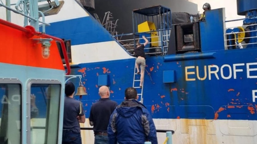 A man walks down a ladder on a blue ship as three men look on with the word 'Euroferry' behind them