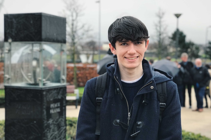 Seamus McLaughlin wears a navy coat and jumper with a black backpack. He smiles at the camera with people in background.