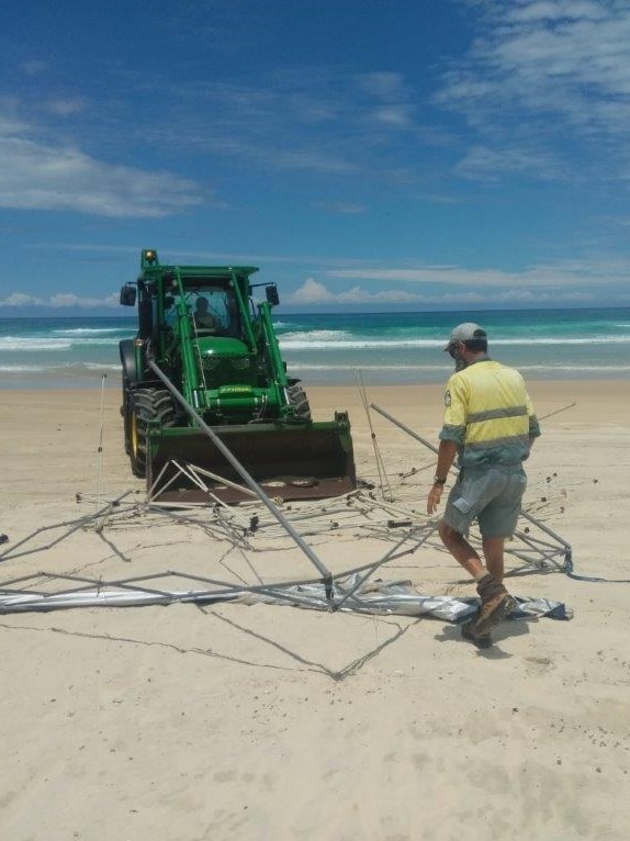 A man in a yellow shirt helping put a metal frame in the bucket of a loader on the beach