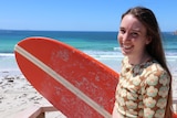 Close up of woman smiling at camera holding orange surfboard with beach and ocean in background