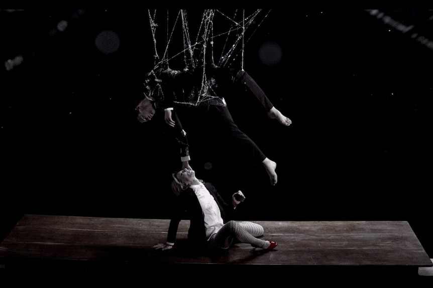 A man in a suit suspended above the ground by a web reaching down to touch a woman's face, who is crouched below him.